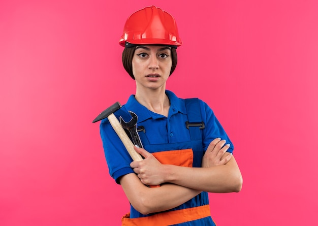 Confident looking at camera young builder woman in uniform holding hammer with open-end wrench crossing hands 