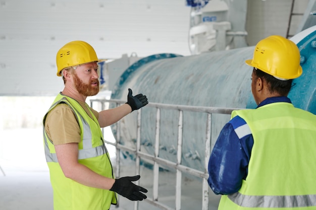 Confident male engineer in uniform gloves and safety helmet talking to colleague