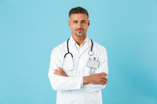 Confident man doctor wearing uniform standing isolated over blue wall, arms folded