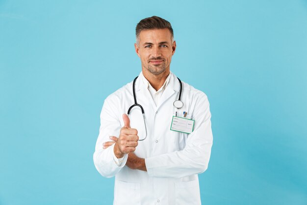 Confident man doctor wearing uniform standing isolated over blue wall, giving thumbs up