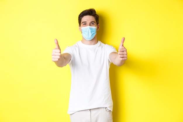 Confident man protecting himself from covid-19 with medical mask, showing thumbs up in approval, yellow wall