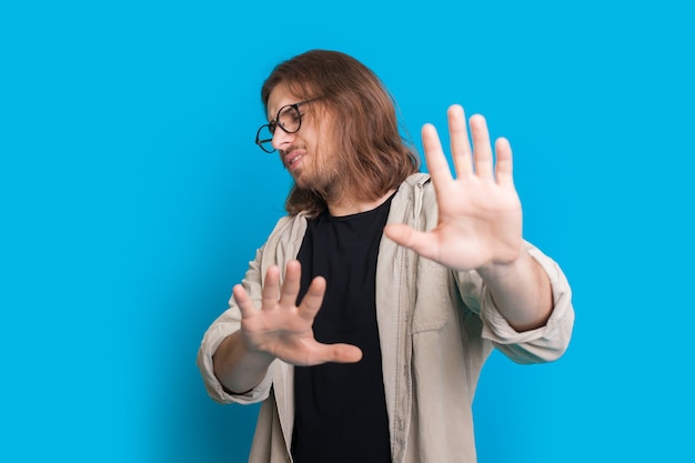 Confident man with long hair and beard is gesturing the refuse sign with palms on a blue studio wall