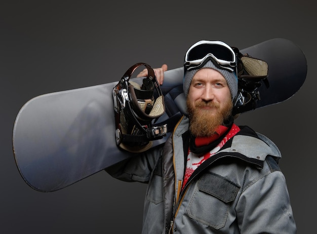 Confident man with a red beard wearing a full equipment holding a snowboard on his shoulder, smiling and looking at a camera, isolated on a dark background.