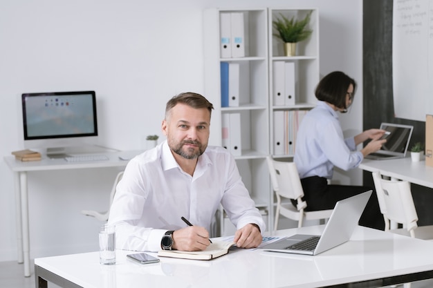 Confident mature businessman looking at you while sitting by desk and writing down working plan with colleague