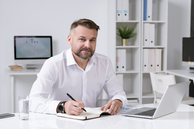 Confident mature businessman sitting by desk in his office while writing down working plan in notebook