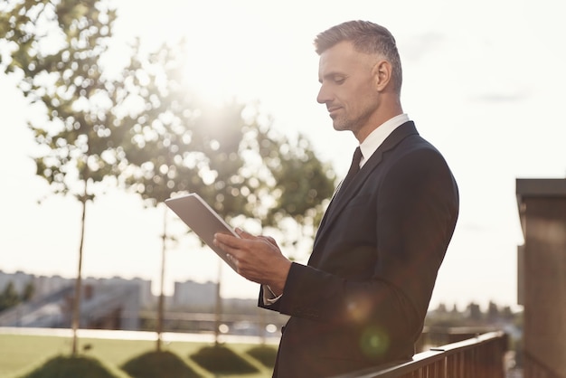 Confident mature businessman working on digital tablet while standing outdoors