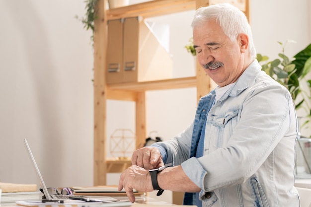 Confident mature man in casualwear pointing at smartwatch on his wrist while checking time before leaving office