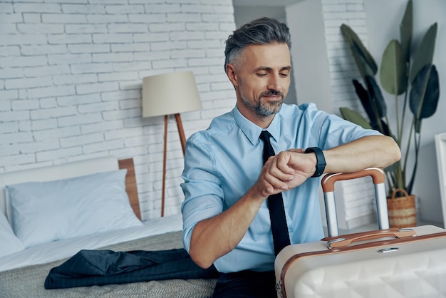 Confident mature man in shirt and tie checking the time while leaning on his luggage at home