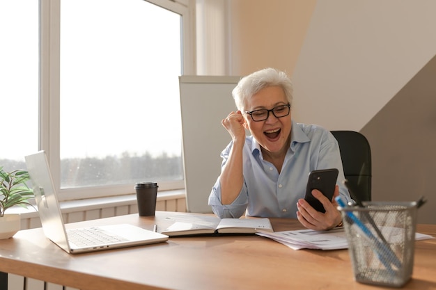 Confident middle aged senior woman euphoric winner with smartphone older mature lady looking at cell