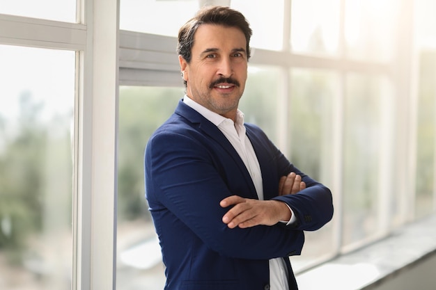 Confident middleaged businessman in a blue suit with crossed arms smiling in a welllit office