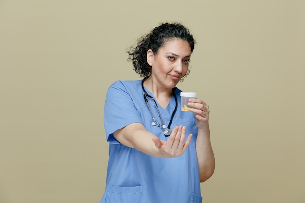 confident middleaged female doctor wearing uniform and stethoscope around neck holding measuring container with pills in it looking at side showing come here gesture isolated on olive background