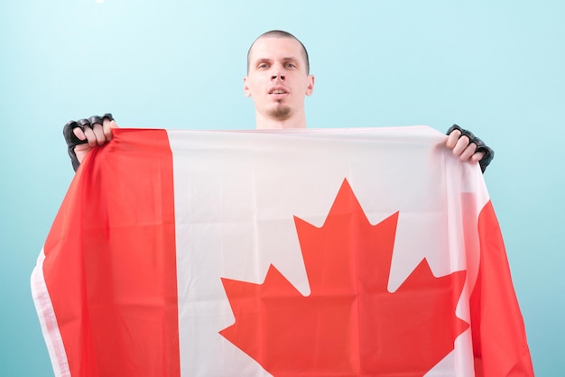 A confident MMA fighter in black gloves holds a Canadian flag on a blue background Pose Knockout Bald Mixed Martial Arts Hit Heavy Tough Hitting Win Goal