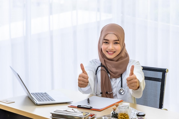 Confident Muslim female woman medical doctor sitting at desk working typing with laptop computer in clinic hospital office with thumb up looking at camera