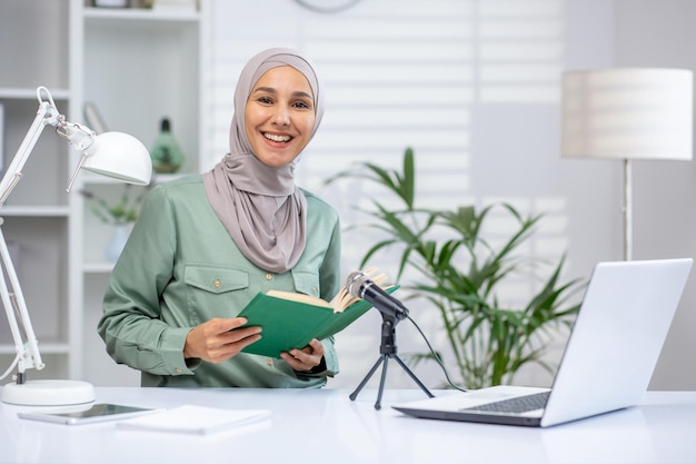 Confident muslim woman in hijab recording an audiobook or podcast while holding a green book in a