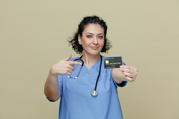 Confident and pleased middleaged female doctor wearing uniform and stethoscope around neck stretching credit card out towards camera pointing at it looking at camera isolated on olive background