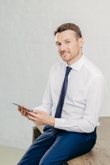 Confident pleased young male entrepreneur in luxury formal clothes reads recieved text message from colleague on modern tablet computer looks joyfully at camera poses in office Vertical shot