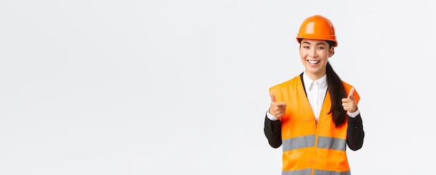 Confident pretty asian female construction manager in safety helmet and reflective jacket pointing fingers at camera praising nice choice good work or well done standing white background