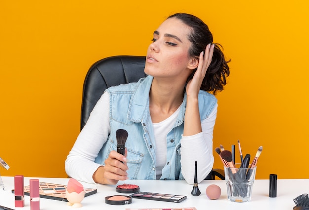 Confident pretty caucasian woman sitting at table with makeup tools holding makeup brush and looking at side isolated on orange wall with copy space