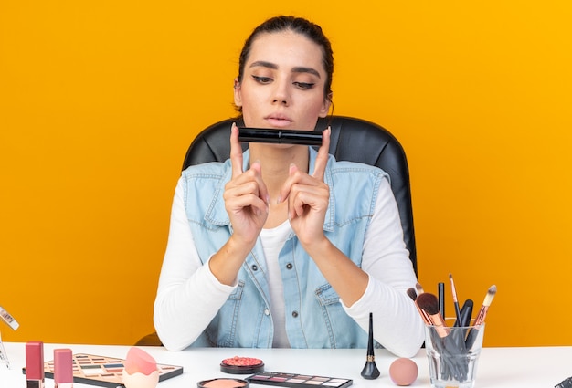 Confident pretty caucasian woman sitting at table with makeup tools holding with two fingers and looking at eyeliner 