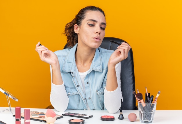 Confident pretty caucasian woman sitting at table with makeup tools looking at eyeliner