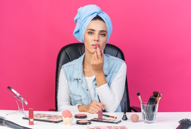 Confident pretty caucasian woman with wrapped hair in towel sitting at table with makeup tools applying lipstick 