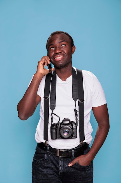 Confident professional photographer having DSLR camera while talking on phone. Smiling heartily young photography enthusiast talking on modern smartphone device while standing on blue background.