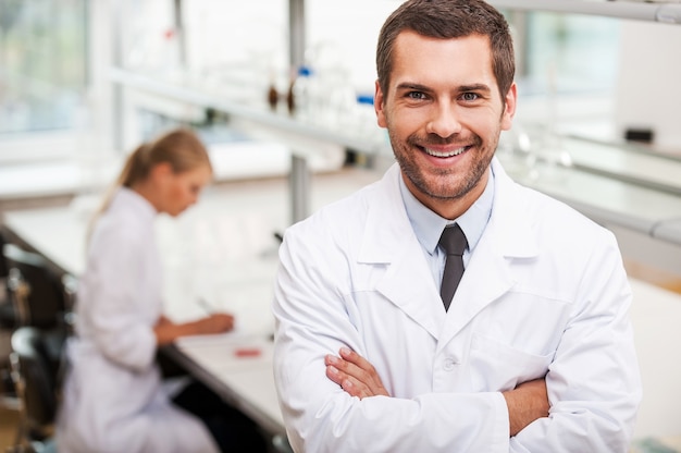 Confident scientist. Happy young male scientist keeping arms crossed and looking at camera while his female colleague working in the background