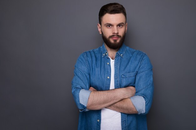 Confident serious hipster modern bearded man in jeans shirt with crossed arms is posing and looking right on the front.