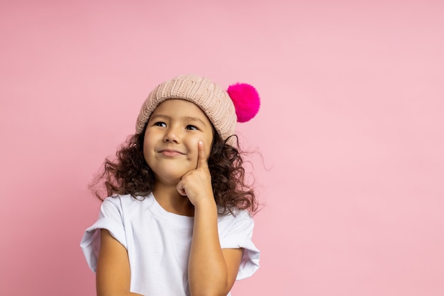 Confident serious little girl, wearing white t shirt, warm hat, keeping forefinger on cheek, looking up with thoughtful expression, thinking, dreaming