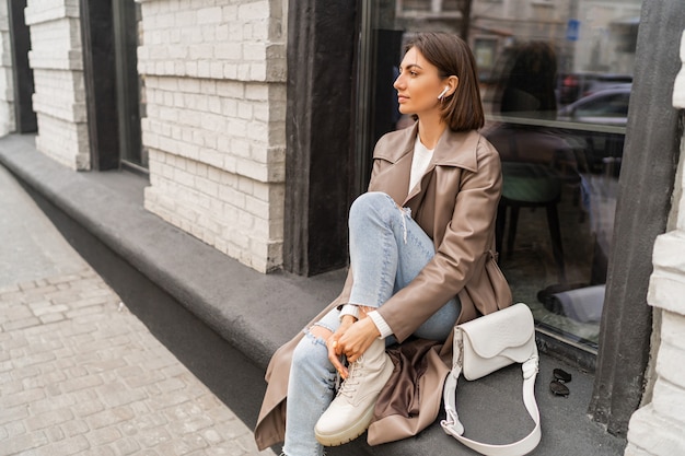 Confident short haired woman wearing trendy beige color coat and white textured leather shoulder bag, walking in street of European city