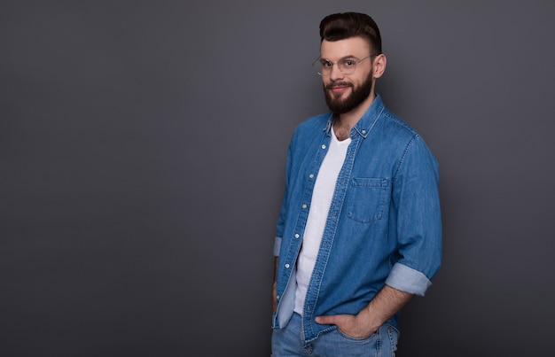 Confident smart modern bearded man in casual denim clothes and eyewear is posing 