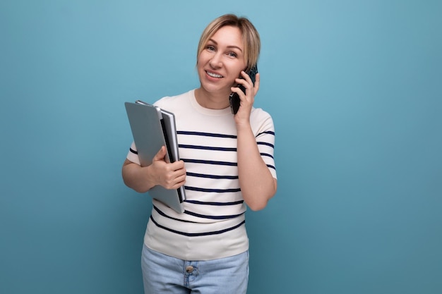 Confident smiling blond girl talking on a smartphone holding a laptop in her hands on a blue