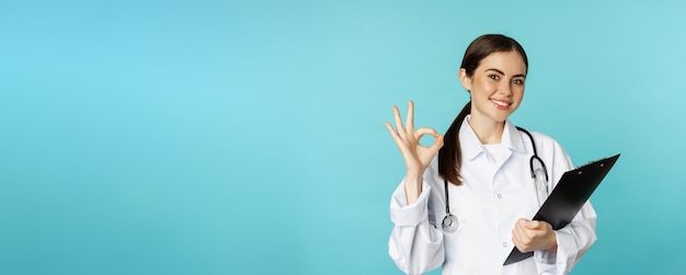 Photo confident smiling doctor woman physician showing okay holding clipboard appointment in hospital stan