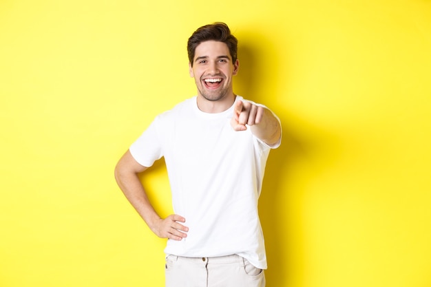 Confident smiling man pointing at you front, standing in white clothes against yellow wall