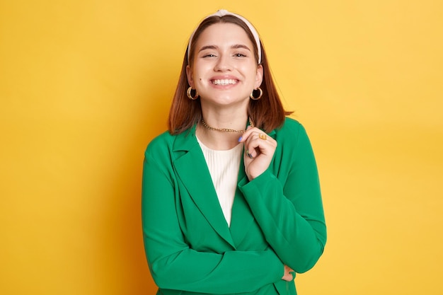 Confident smiling woman wearing green jacket posing isolated over yellow background looking at camera with toothy smile rejoicing having satisfied face