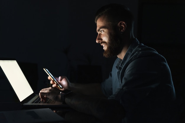 Confident smiling young man working on laptop computer while sitting at the table indoors at night, using mobile phone