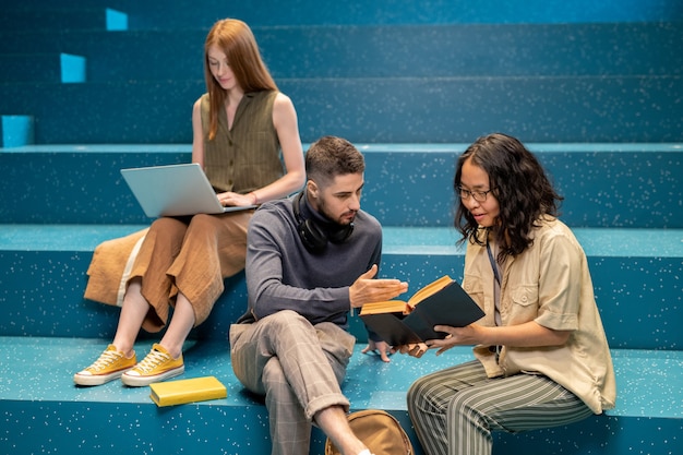 Confident student explaining something to female groupmate while pointing at page of book