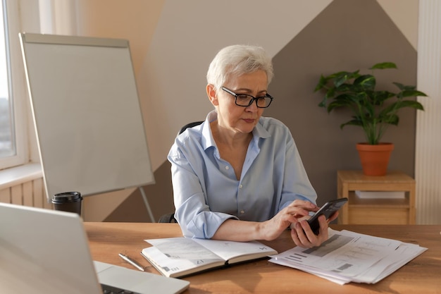 Confident stylish european middle aged senior woman using smartphone at workplace stylish older matu