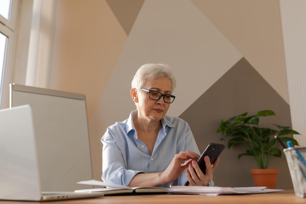 Confident stylish european middle aged senior woman using smartphone at workplace stylish older matu