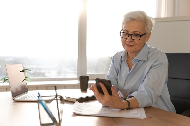 Confident stylish european middle aged senior woman using smartphone at workplace stylish older matu