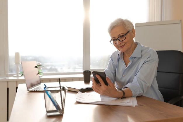 Confident stylish european middle aged senior woman using smartphone at workplace stylish older matu