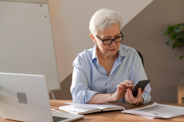 Confident stylish european middle aged senior woman using smartphone at workplace stylish older matu