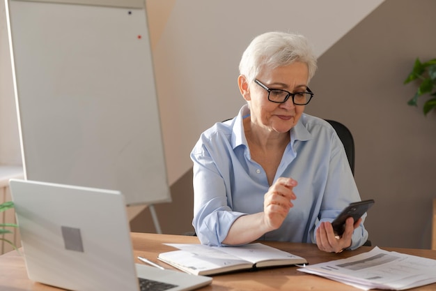 Confident stylish european middle aged senior woman using smartphone at workplace stylish older matu