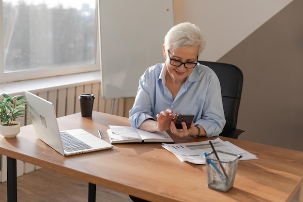 Confident stylish european middle aged senior woman using smartphone at workplace Stylish older mature 60s gray haired lady businesswoman with cell phone in office Boss leader using internet apps