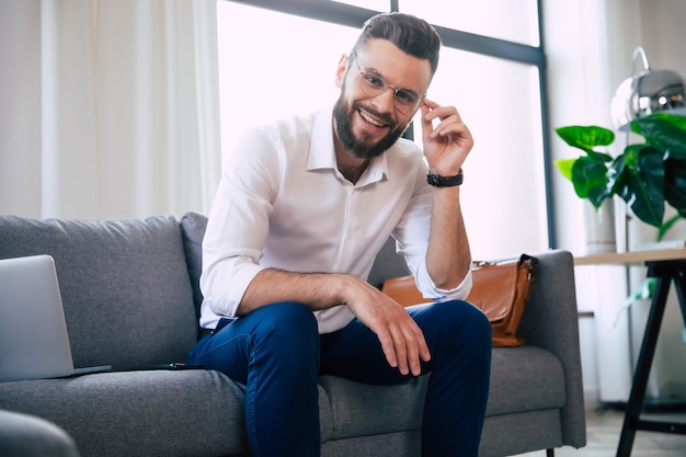 Confident stylish handsome bearded business man in glasses and formal smart wear is sitting on the couch in the modern office or room