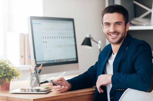 Confident and successful. Confident young man holding smart phone and smiling while sitting at his working place in office