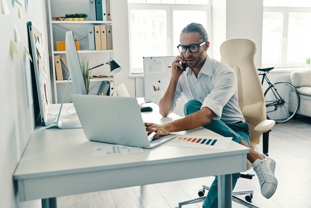 Confident and successful. Good looking young man in shirt talking on the smart phone while sitting in the office