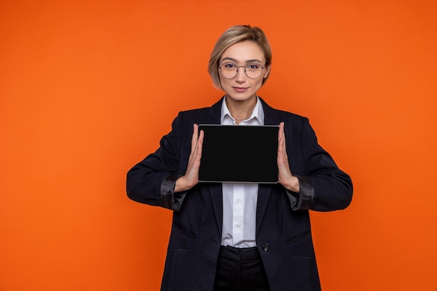 Confident woman wearing black official style suit showing blank screen of tablet isolated over orange background