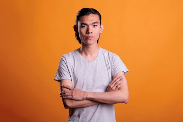 Confident young adult asian man standing with arms crossed, looking at camera, front view. Teenager with serious facial expression portrait, attractive person medium shot