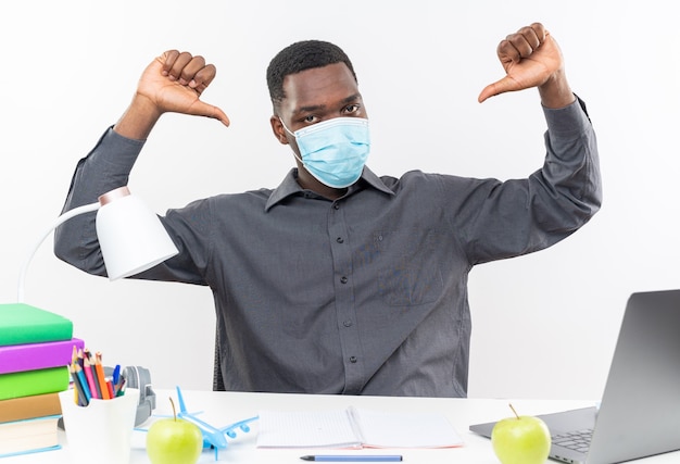 Confident young afro-american student wearing medical mask sitting at desk with school tools pointing at himself 